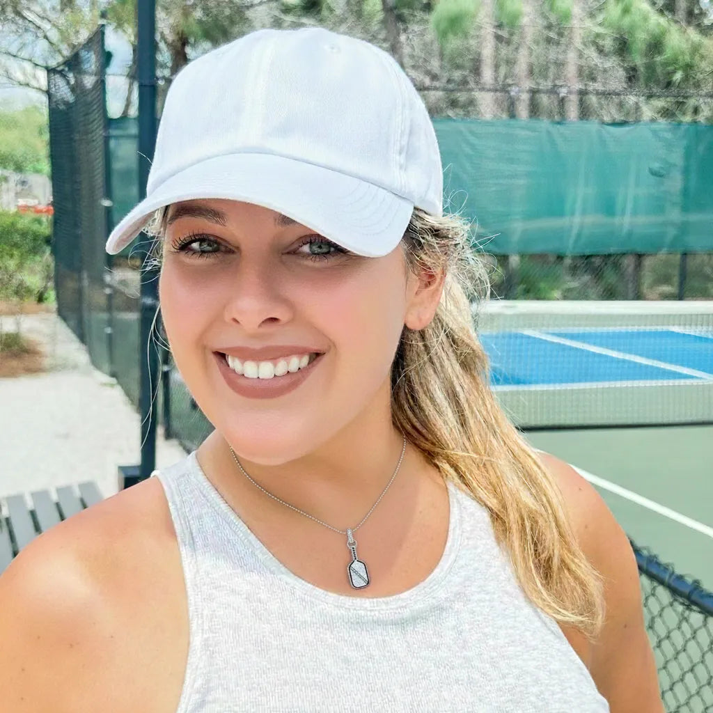 Woman wearing a white cap, tank top and silver pickleball necklace, standing on a tennis court