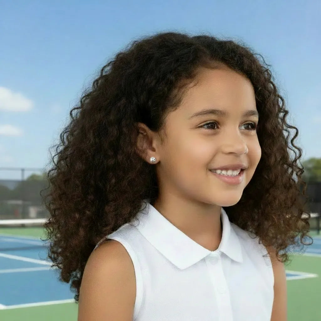 Young girl with curly hair wearing a white sleeveless top and silver pickleball stud earring on a sports court.