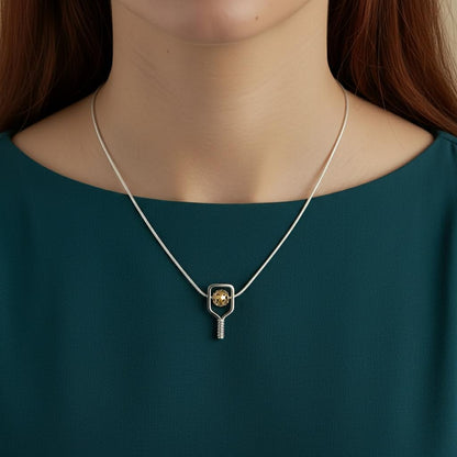 Necklace with a pickleball paddle and ball pendant worn by a person wearing a teal shirt.