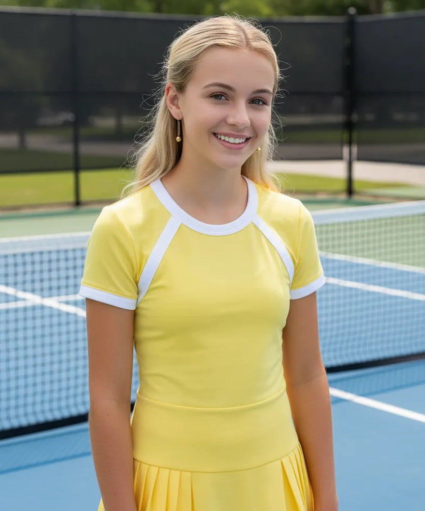 Young teenager in a yellow dress wearing gold pickleball dangle earrings standing on a tennis court