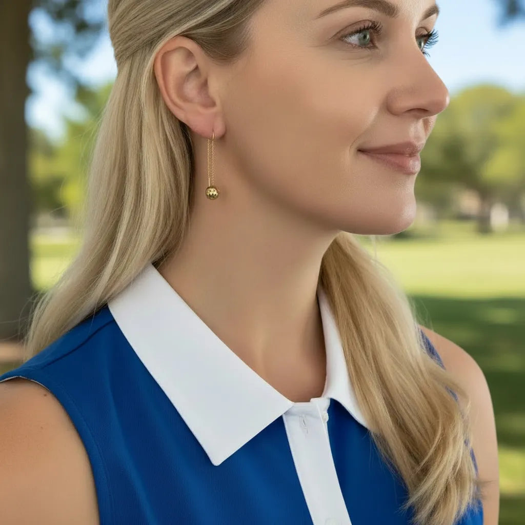 Woman wearing a blue sleeveless top and gold dangle pickleball earrings with a white collar in an outdoor setting
